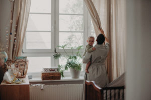 Eine Mutter öffnet die Vorhänge mit ihrem Baby auf dem Arm entstanden bei dokumentarischem Familienfotoshooting in Freiburg